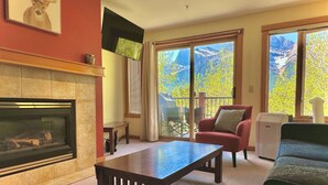 A living room with a fireplace, a sofa, a coffee table, and a view of mountains through large windows. - C303 Peaceful Canmore Corner- Downtown, Free parking, View, 2BR, 2BT, Sleeps 7 (Canmore)