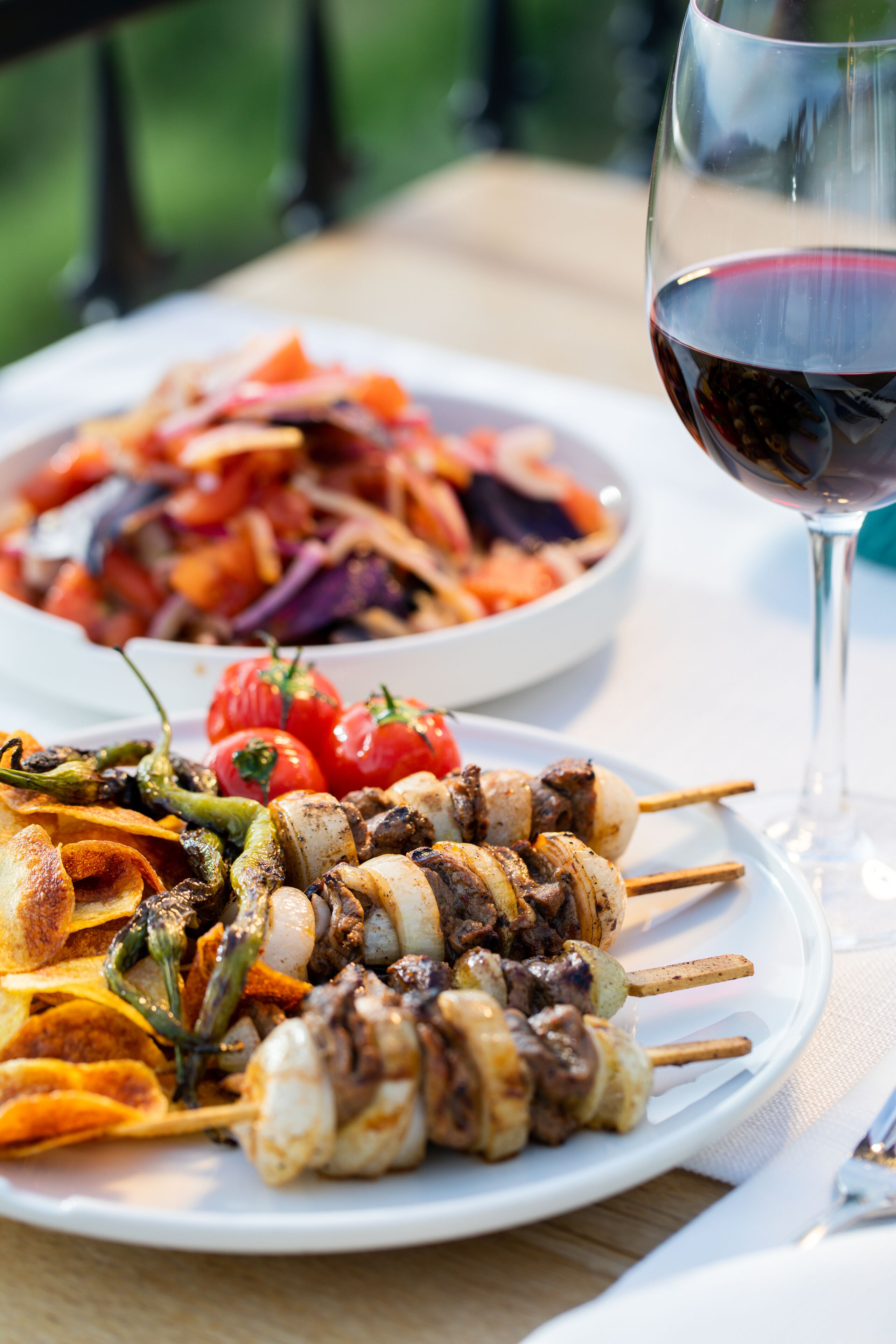 Un plato con brochetas de carne y verduras, una copa de vino tinto y un bol de ensalada.