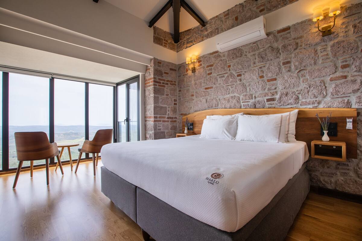 A bedroom with a large bed, a stone wall, a wooden headboard, and a view through a large window.