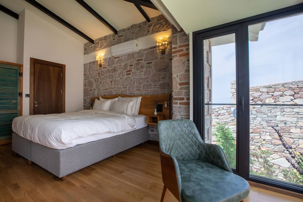 A bedroom with a bed, a chair, a stone wall, and a view through a glass door.