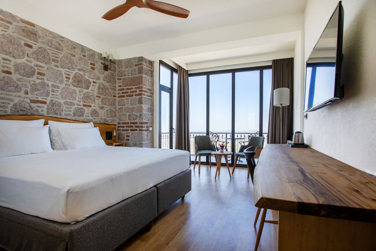 A hotel room with a large bed, a wooden desk, a balcony with a view, and a stone wall.