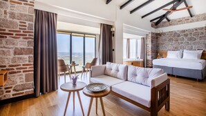 Una habitación de hotel moderna con una cama grande, un sofá, una zona de comedor con mesa y sillas, y vistas del paisaje a través de amplios ventanales.