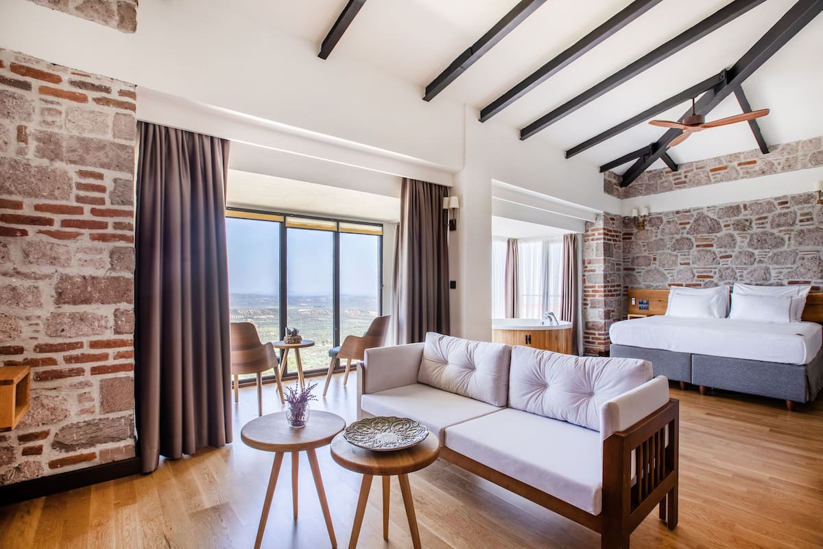 A modern hotel room with a large bed, a sofa, a dining area with a table and chairs, and a view of the landscape through large windows.