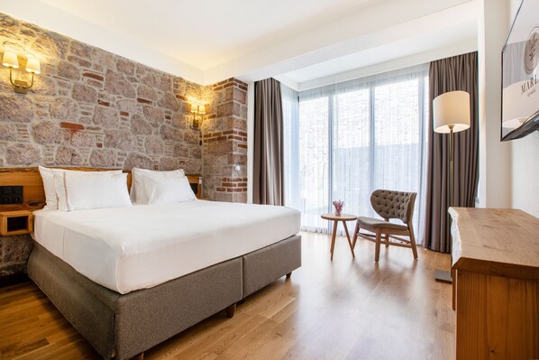 A hotel room with a large bed, a stone accent wall, a wooden nightstand, a small round table with a chair, and a large window with curtains. - Marlo Ayvalik Adult Only +12 (Ayvalik)
