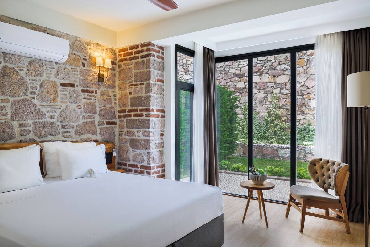 A hotel room with a stone wall, a large bed, a chair, and a view of greenery outside.