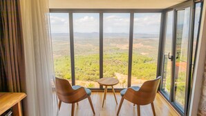 Una habitación con un ventanal que ofrece vistas a un paisaje. Hay dos sillas de madera y una mesita en la habitación.