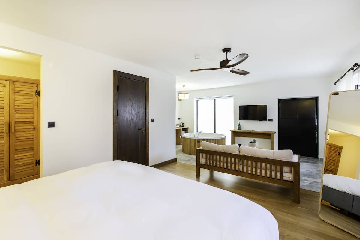 A hotel room with a large bed, a wooden bench, a round table, a mirror, and a ceiling fan.