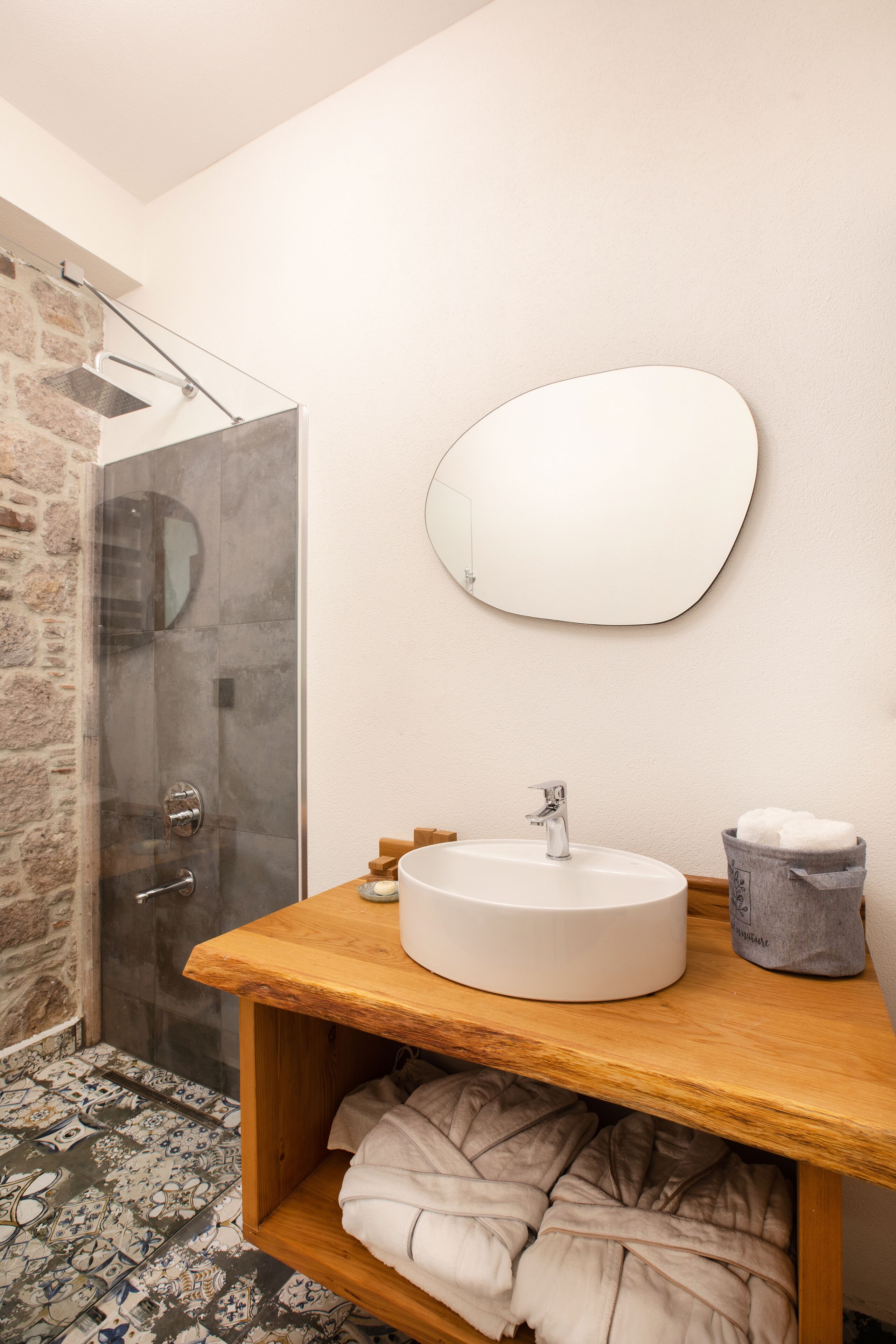 A modern bathroom with a stone shower, a wooden vanity, a round mirror, and a white sink.