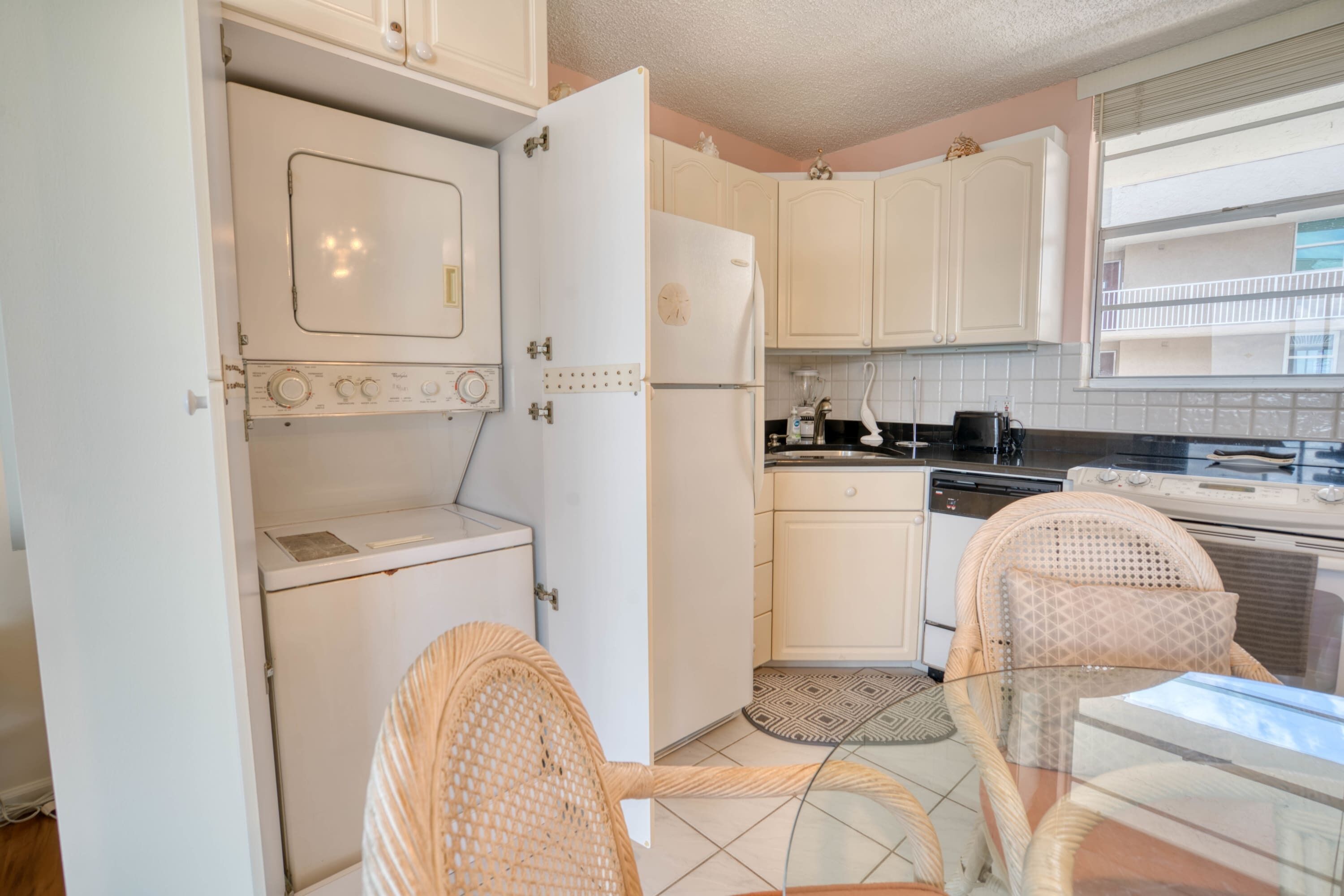 Cocina compacta con armarios blancos, un refrigerador, una estufa y un comedor con sillas de mimbre y una mesa de vidrio.