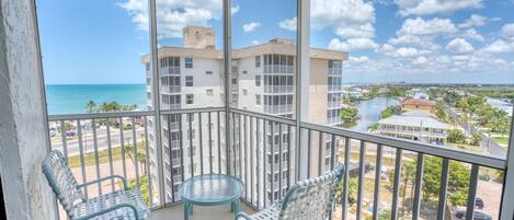 A balcony with two wicker chairs and a small round table, overlooking a beach and a cityscape with buildings and a river.