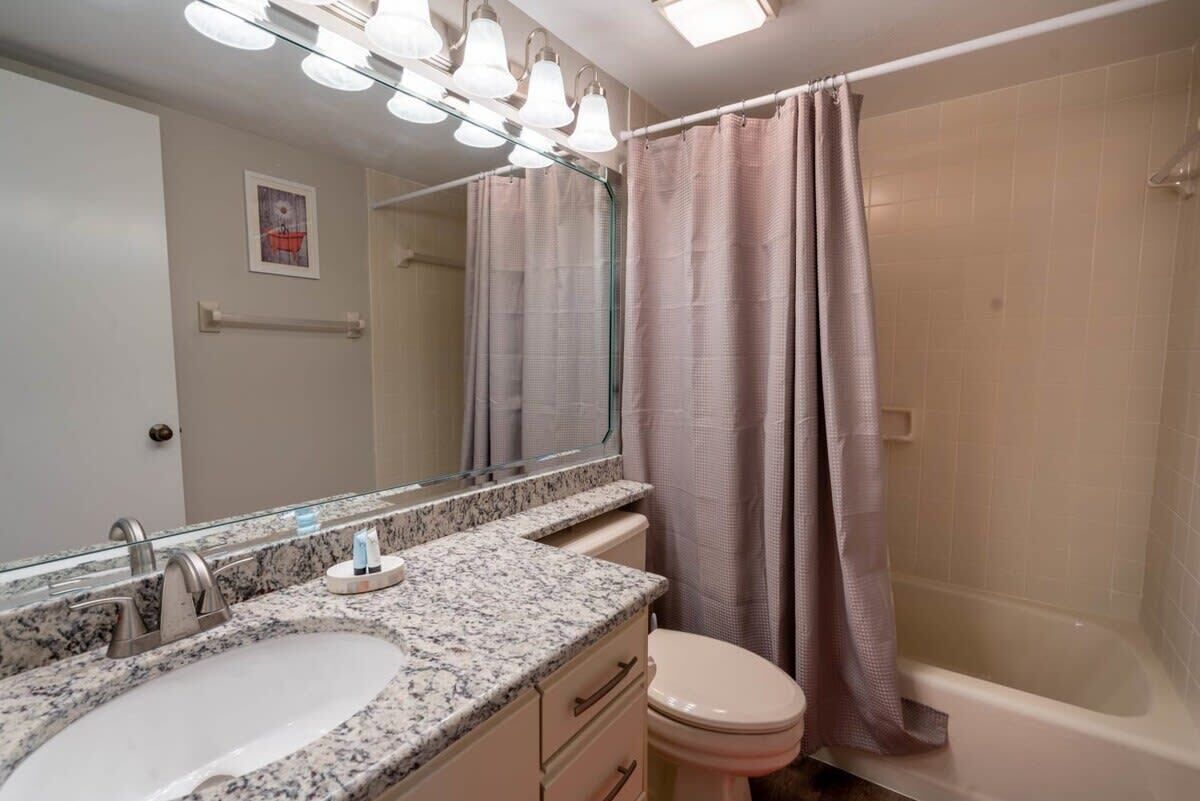 Baño con lavamanos de granito, espejo con iluminación, inodoro y ducha con cortina rosa.
