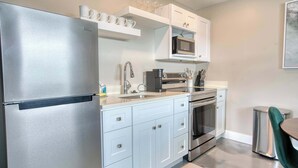A modern kitchen with stainless steel appliances, white cabinetry, and a small dining area with a green chair. - Mango Street Inn Suite 5 (Fort Myers Beach)