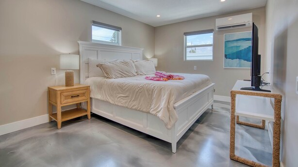 A bedroom with a white bed, a wooden nightstand, a woven side table, and a wall-mounted TV. - Mango Street Inn Suite 5 (Fort Myers Beach)