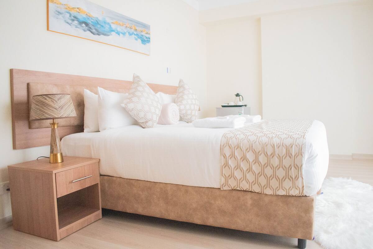 Un dormitorio ordenado con cama, mesita de noche, lámpara y un cuadro en la pared.