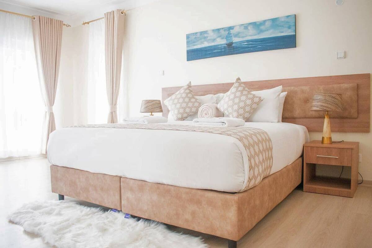 Un dormitorio con una cama grande, un cabecero de madera, dos mesitas de noche con lámparas y un cuadro de un velero en la pared.