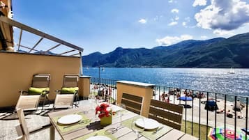 Una terrazza con tavolo apparecchiato per mangiare, vista su una spiaggia e un lago con barche.