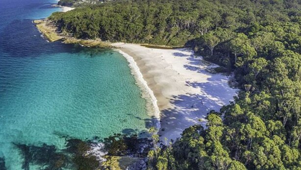 An aerial view of a coastal area with a sandy beach, clear turquoise waters, and dense green forest.