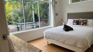 A bedroom with a large window, a bed with a white duvet, and a view of an outdoor area with a table and chairs.
