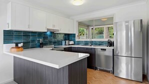 A modern kitchen with white cabinets, a central island, a stainless steel refrigerator, and a window with a view of greenery. - Our Beach House by Experience Jervis Bay (Vincentia)