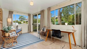 A spacious room with a large window, a desk, a chair, a sofa, and a striped rug. - Our Beach House by Experience Jervis Bay (Vincentia)