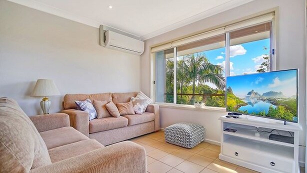 A living room with a beige sofa, a TV on a white stand, and a window with a view of greenery and a lake.
