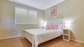 A bedroom with a bed, a nightstand, a lamp, a window with blinds, and a picture on the wall.
