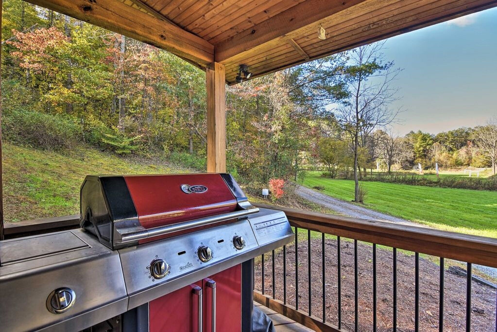 A covered outdoor area with a barbecue grill, wooden deck, and a view of a landscaped garden and trees.