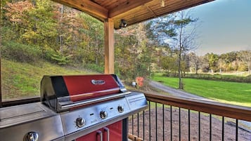 A covered outdoor area with a barbecue grill, wooden deck, and a view of a landscaped garden and trees.