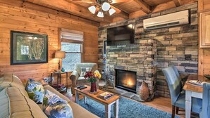 A cozy cabin interior with a stone fireplace, wooden walls, a ceiling fan, and a comfortable seating area.