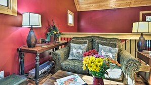 A cozy room with a wooden ceiling, a red wall, a patterned sofa with cushions, a small table with a lamp, and a vase of flowers.