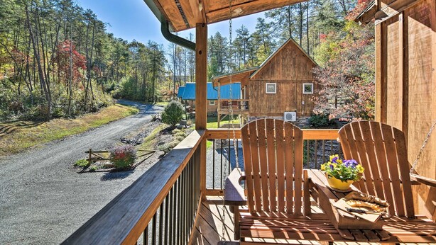 A wooden deck with two Adirondack chairs, a small table with a bowl of flowers, and a view of a forested road.