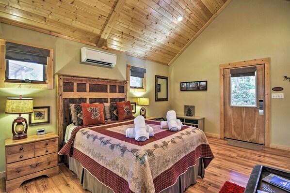 Un dormitorio acogedor con techo de madera, una cama grande con colcha, dos lámparas de noche, una mesita de noche y una puerta con vistas a árboles.