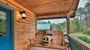 A wooden porch with two Adirondack chairs, a small table with flowers, and a view of a house with a green roof. - Dog-Friendly Weaverville Cabin King Bed,Hot Tub&FP (Weaverville)