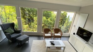 Una sala de estar moderna con un sofá gris, un sillón negro y dos sillas de mimbre. Un televisor de pantalla plana está sobre un mueble blanco, y hay una mesa de centro con un control remoto. Amplias puertas de cristal dan a un exuberante jardín verde.