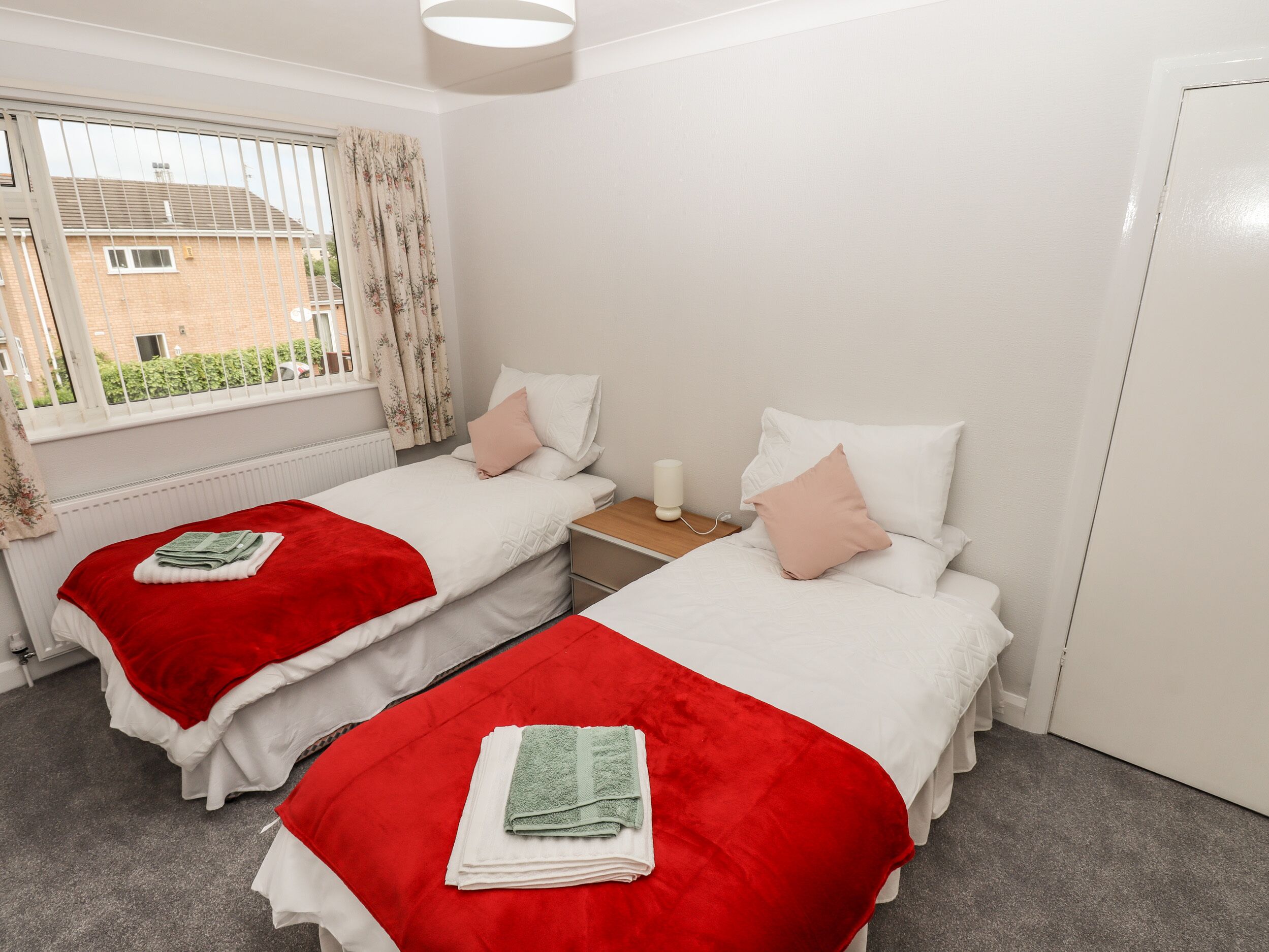 Una camera matrimoniale con due letti, ciascuno con coperta e cuscini rossi, un comodino con una lampada e una finestra con tende.