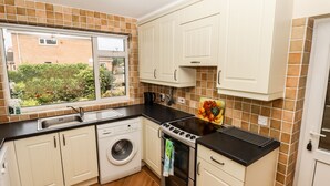 A compact kitchen with white cabinets, a built-in washing machine, a stove, and a window overlooking a garden.