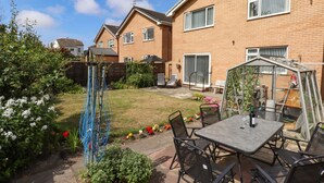 A residential backyard with a patio table and chairs, a garden with various plants, and a brick house in the background.