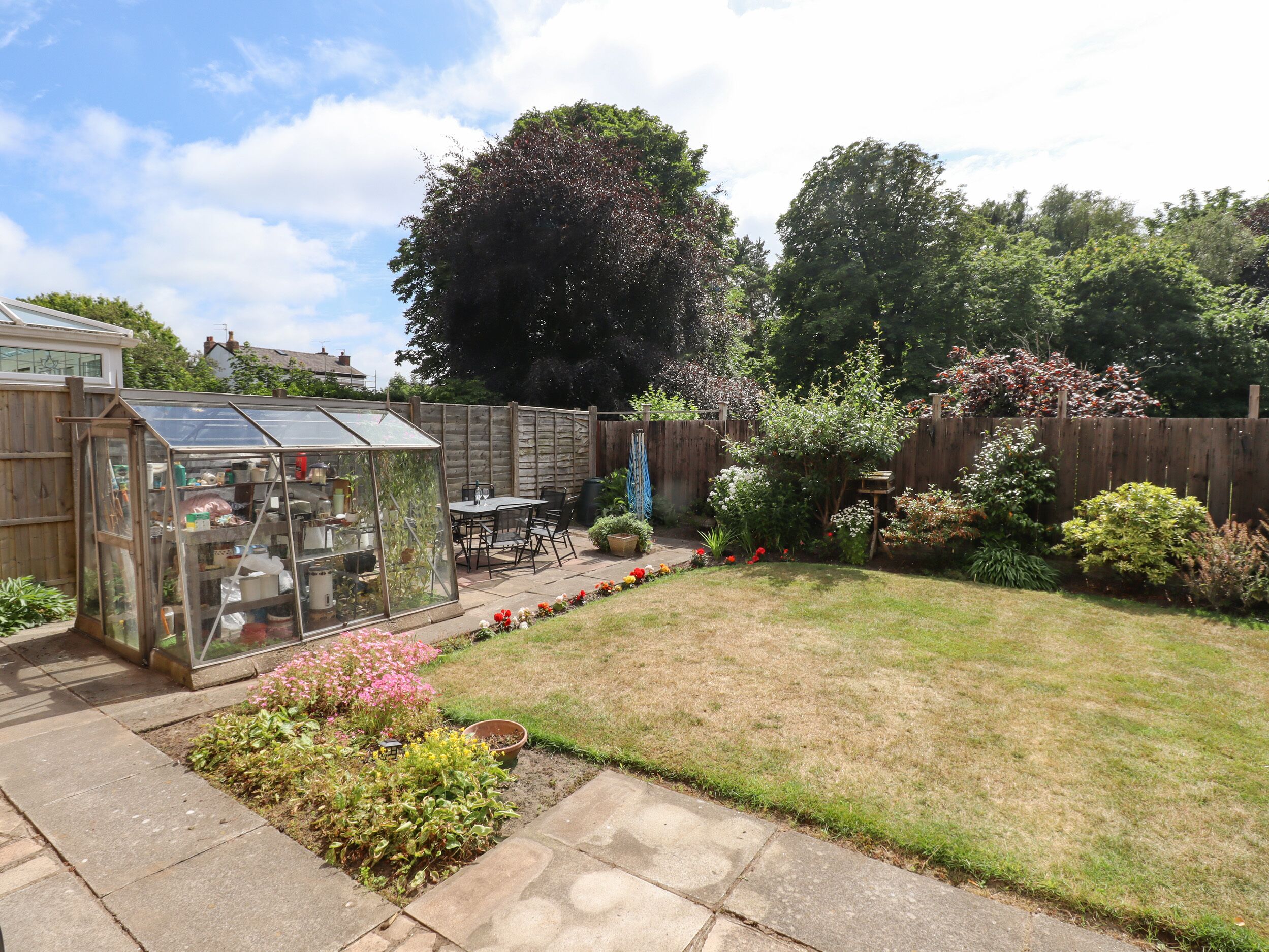 A backyard with a glass greenhouse, a patio table, and various plants.