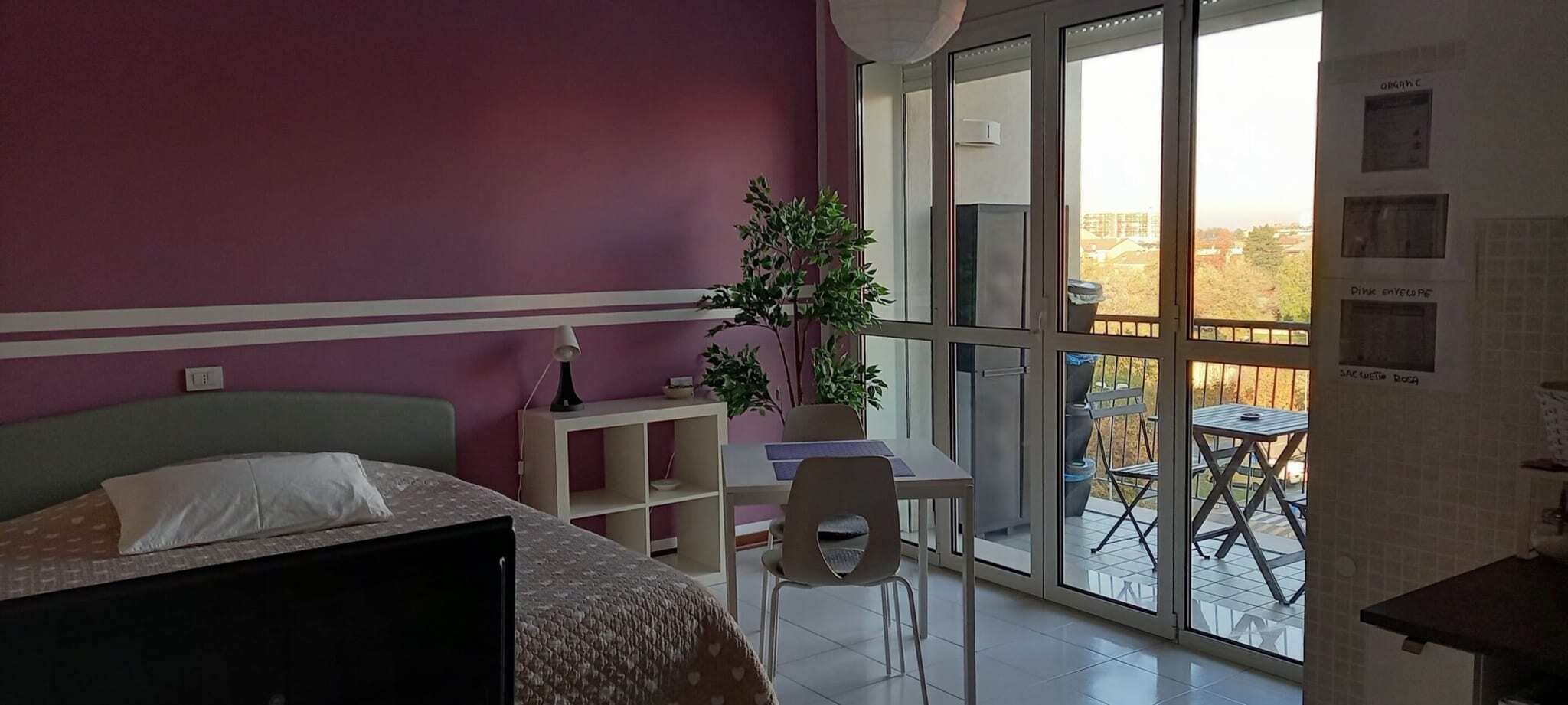 Ein Zimmer mit Bett, Schreibtisch, Stuhl, einer Pflanze und einem Balkon mit Tisch und Stühlen.