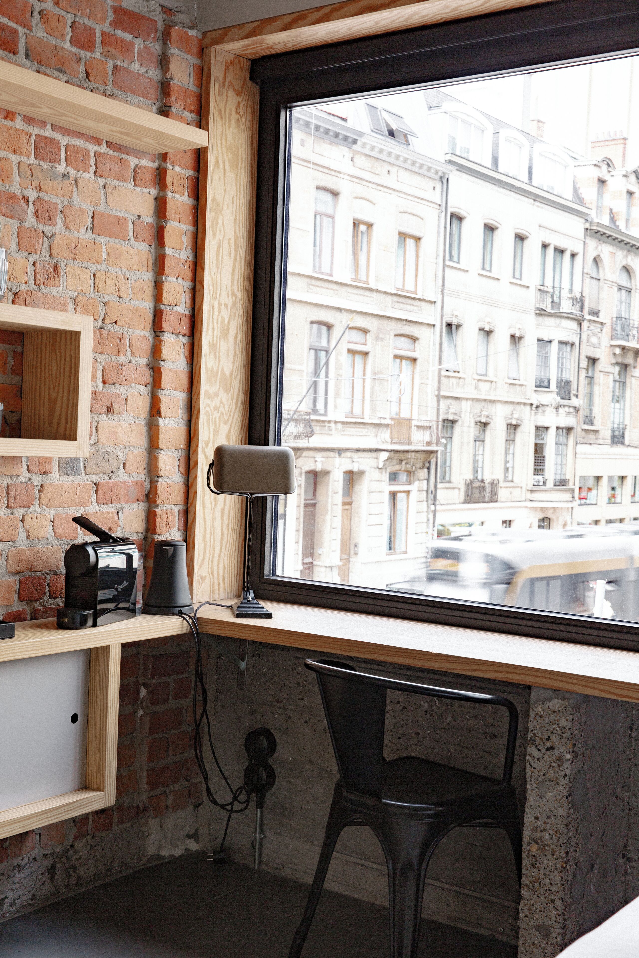 A room with a brick wall, a wooden shelf, a desk with a chair, a window with a view of buildings, and a lamp.