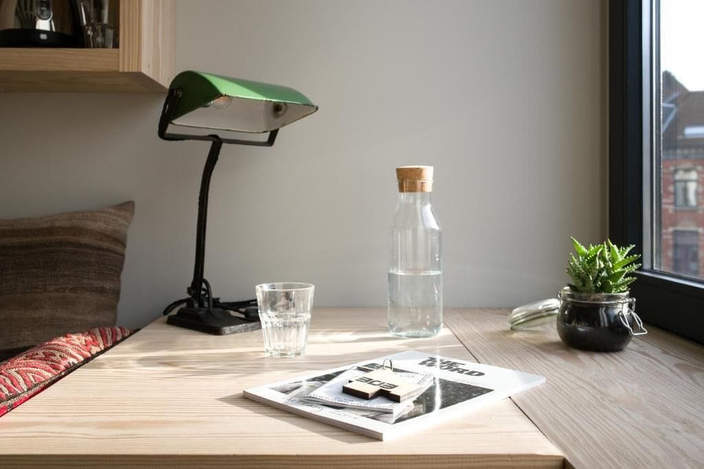 Een houten tafel met een groene bureaulamp, een glas water, een waterfles, een kleine potplant en een tijdschrift.