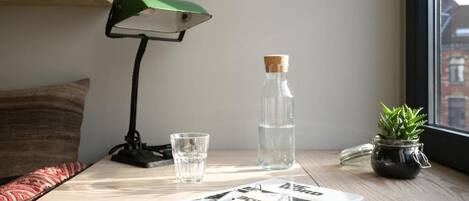 A wooden table with a green desk lamp, a glass of water, a bottle of water, a small potted plant, and a magazine.