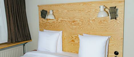 Una habitación de hotel con cabecera de madera, dos lámparas de noche, una cama con ropa blanca, una mesita de noche de madera pequeña y una ventana con cortinas.