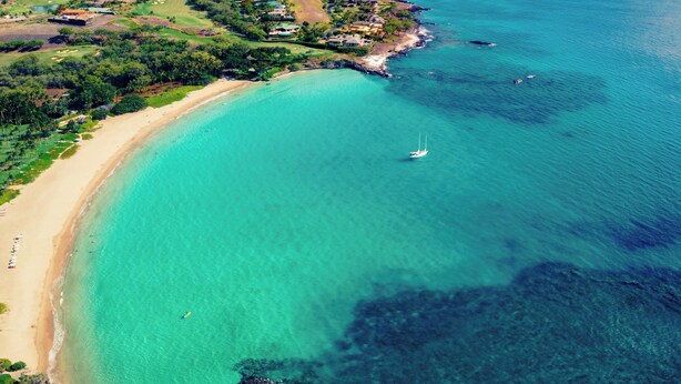 Vue aérienne d’une zone côtière avec une plage, des eaux turquoise et limpides, et un petit voilier.