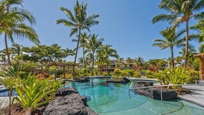 Une piscine d’un complexe hôtelier, avec une eau cristalline, entourée de palmiers et de plantes tropicales.