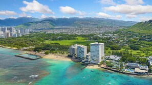 A coastal cityscape with a beach, clear turquoise waters, and a backdrop of mountains.