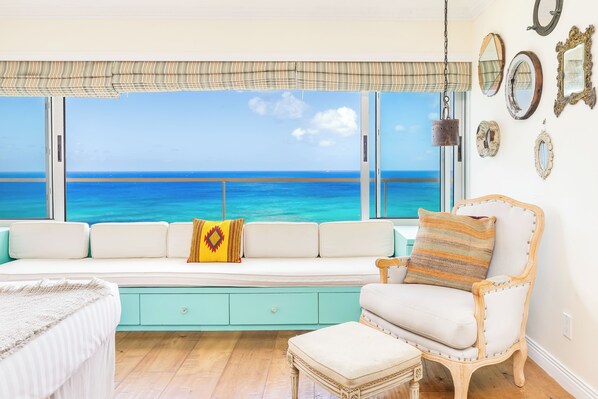 A room with a view of the ocean, a white sofa with cushions, a wooden chair, and a window with a striped valance.