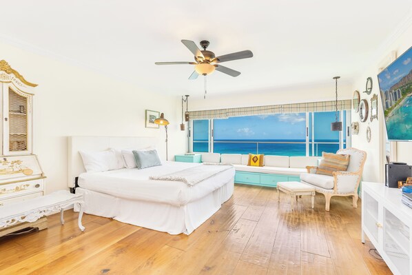 A bedroom with a bed, a sofa, a ceiling fan, and a large window with a view of the ocean.