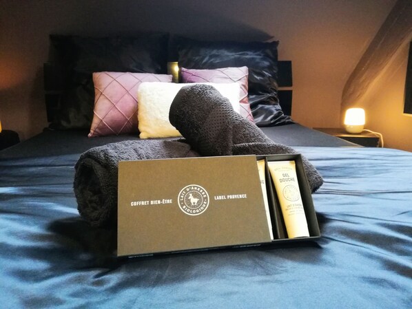Una cama bien tendida con una toalla enrollada, una almohada y una caja etiquetada como "COFFRET BIEN-ÊTRE" sobre una colcha azul.