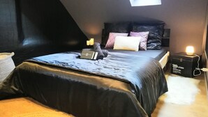 Een slaapkamer met een schuine kap, een dakraam, een bed met een zwarte matras en kussens, een nachtkastje met een lamp, en een doos met de tekst '20 Liter' op de vloer.